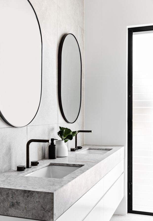 A refined contemporary bathroom vanity with a thick pale grey stone benchtop, two integrated rectangular basins, matte black wall-mounted taps and fixtures, and white built-in cabinetry. Large rounded-rectangle mirrors with slim black frames sit against a softly textured light wall, while a black-framed window with white shutters provides natural light.