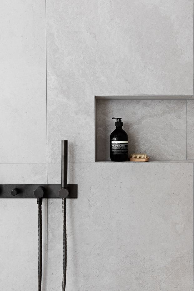 Close-up of a minimalist contemporary shower wall clad in large pale grey textured tiles or stone-look panels. A recessed wall niche contains a black pump bottle and wooden brush, while a matte black shower control bar, handheld shower hose, and vertical rail provide strong dark accents.