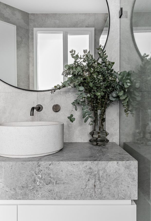 A refined bathroom vanity featuring a round white vessel basin on a substantial grey stone benchtop, with a wall-mounted dark tap and matching control. Large frameless round mirrors, pale grey stone-look wall tiles, white cabinetry, and a glass shower enclosure create a calm, monochromatic setting. A tall glass vase of eucalyptus adds an organic accent.
