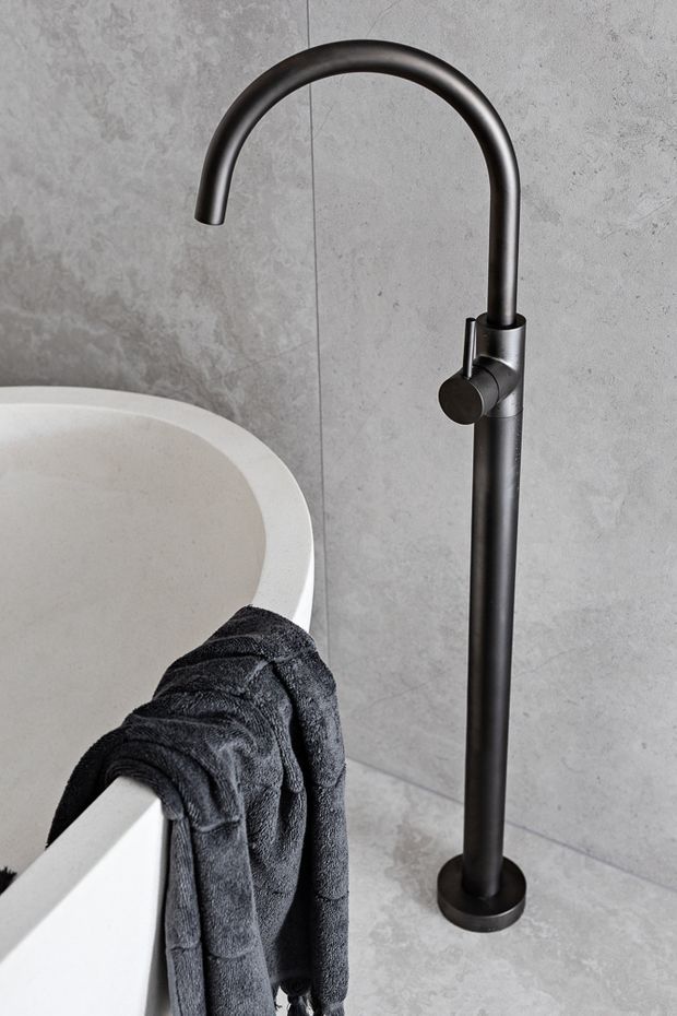 Close-up product-style photograph of a freestanding matte black bath faucet beside a white freestanding bathtub. The fixture has a tall cylindrical body, curved gooseneck spout, and single side control, set against large grey concrete-look wall and floor tiles. A dark towel drapes over the tub edge.