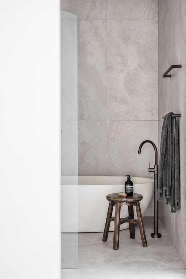 A minimalist contemporary bathroom viewed through a partially open doorway, featuring large pale grey stone-look wall and floor tiles, a freestanding white bathtub, matte black freestanding bath filler, black wall-mounted towel hooks, and a dark towel. A rustic wooden stool with a black soap dispenser sits beside the tub.