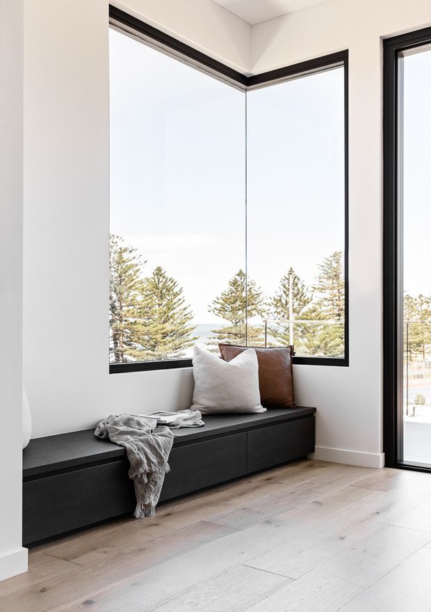 Bright, minimalist interior corner featuring a built-in dark timber window seat with integrated cabinetry, a small selection of cushions and a throw, pale timber flooring, white walls, and expansive black-framed corner glazing overlooking trees and water. A full-height glazed door is visible on the right.