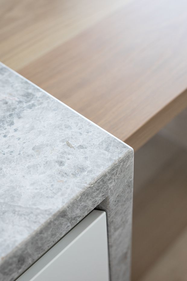 Close-up detail of a pale grey, heavily veined natural stone benchtop with a rounded edge, adjoining warm light-wood cabinetry or paneling and a flush light-grey panel.