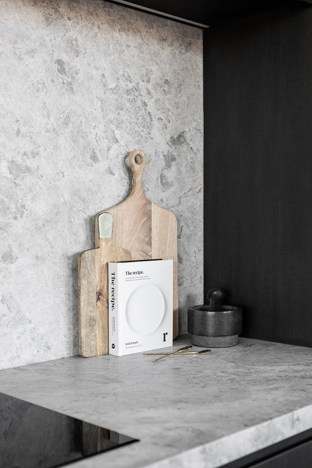 A close-up vignette of a refined kitchen or pantry benchtop featuring a grey, heavily veined stone surface and matching stone backsplash. Dark textured cabinetry frames the scene, with timber chopping boards, a cookbook, ceramic vessel, and metal utensils arranged as styling elements. The palette is predominantly grey, black, and light timber with a moody, contemporary feel.