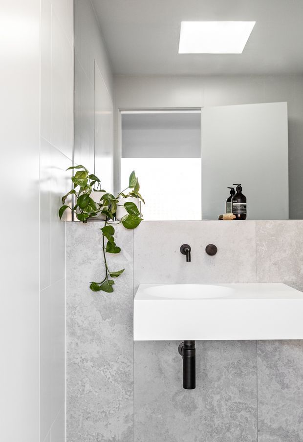 A bright, compact contemporary bathroom featuring a wall-mounted white rectangular basin, black tapware and exposed black waste pipe, large light-grey stone-look wall tiles, a frameless mirror, a skylight, and a trailing green plant on the vanity ledge.