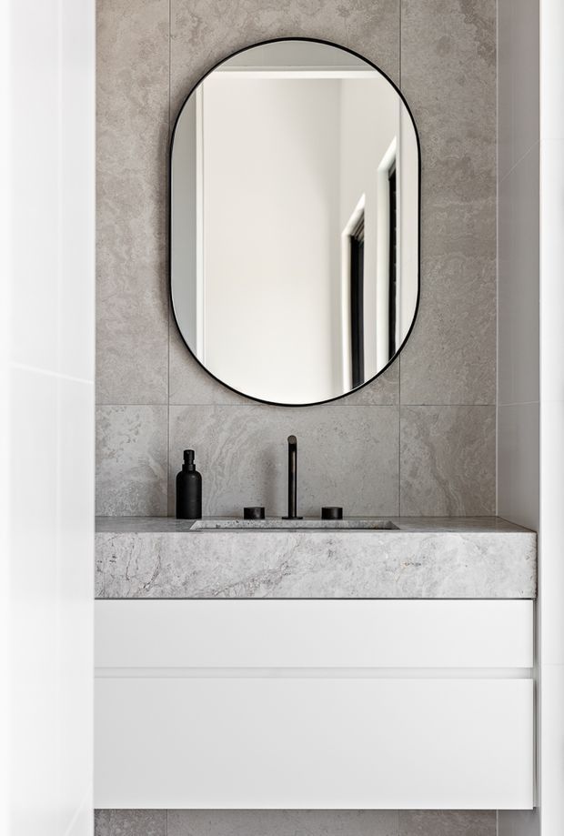 A compact, modern bathroom vanity with a white floating two-drawer cabinet, pale grey stone-look tiled wall, and a matching grey stone benchtop with integrated basin. A black wall-mounted or deck-mounted tap, black handles, and a rounded rectangular black-framed mirror provide contrast. Minimal monochrome styling and clean lines create a refined contemporary appearance.