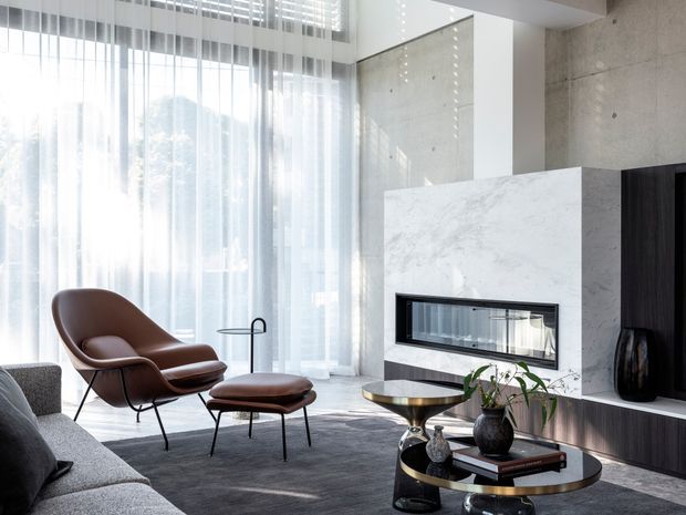 Bright, refined contemporary living room with floor-to-ceiling sheer-curtained glazing, a sculptural fireplace clad in pale veined stone, dark timber built-in cabinetry, and a grey sectional sofa. A leather lounge chair and ottoman sit on a dark rug beside layered round coffee tables with plants and books.