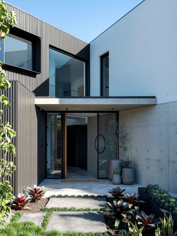 Contemporary multi-level residential facade viewed from a landscaped entry path. The home combines dark vertically ribbed cladding, smooth pale concrete walls, expansive black-framed glazing, and a projecting concrete upper-level slab. A recessed glass entry sits beneath the cantilever, with tropical planting and large concrete stepping slabs in the foreground.