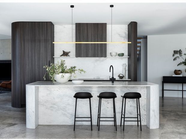 Contemporary open-plan kitchen centred on a large white marble-look island with three black bar stools. Dark timber cabinetry contrasts with pale stone wall cladding and benchtops, while a slim linear pendant light spans the island. Polished concrete flooring and minimal black-framed furniture create a refined, minimalist interior.
