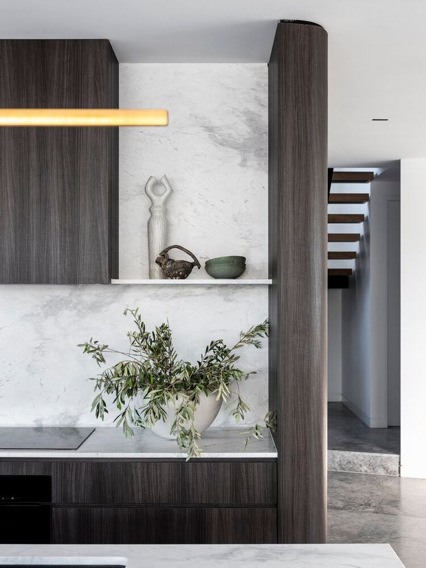 A refined contemporary kitchen interior featuring dark vertically grained timber cabinetry, a pale veined stone backsplash and benchtop, open display shelving with sculptural ceramics, and a large curved-edge cabinetry panel framing a hallway. A linear pendant light and olive branch arrangement add restrained warmth. The adjacent passage includes dark stone flooring and floating timber stair treads.
