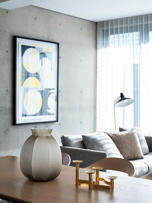 A refined contemporary living room with a raw exposed concrete feature wall, large sheer-curtained window, grey sectional sofa, and a timber coffee table in the foreground. Sculptural neutral-toned decor, an abstract geometric artwork, and a brass candleholder add restrained accents to the bright, softly lit space.