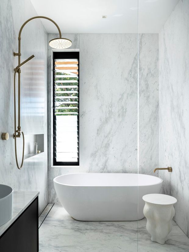 A bright, refined bathroom clad in pale veined marble-effect surfaces, featuring a freestanding white bathtub, frameless glass shower enclosure with a brushed brass shower set, wall-mounted bath filler, a narrow black-framed louvred window, and dark vanity cabinetry at the left edge. A sculptural white stool adds a soft organic accent.