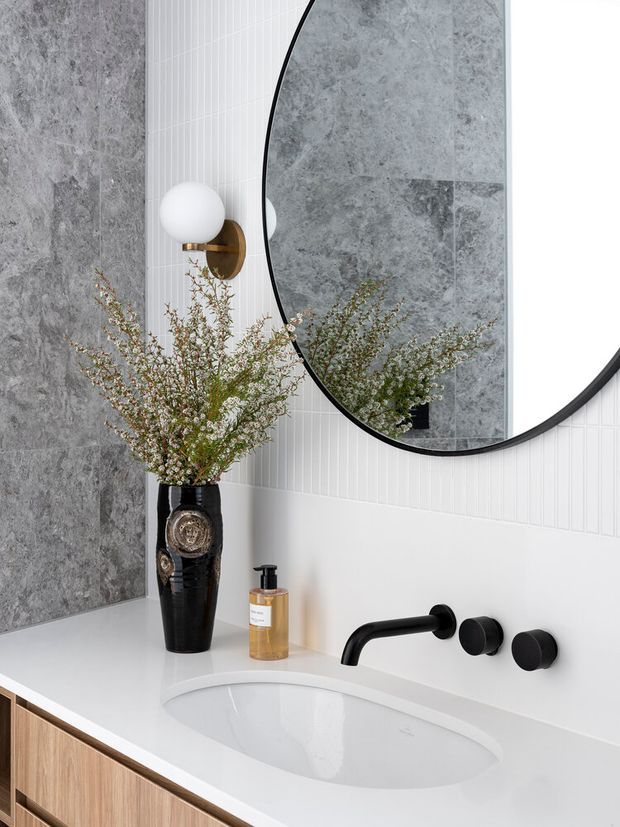 A refined contemporary bathroom vanity featuring a white integrated basin, floating light-wood cabinetry, matte black wall-mounted tapware, grey stone-look wall tiles, and vertically ribbed white wall tiles. A large curved black-framed mirror reflects the basin area, while a sculptural wall sconce and decorative vase add warmth and character.