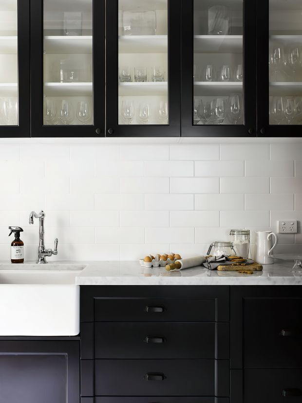 A close-up view of a refined kitchen featuring black framed glass-front upper cabinetry displaying glassware, white subway-tile splashback, a pale stone benchtop, black lower drawers and cabinetry, and a white farmhouse-style sink with polished metal tapware. Small countertop items include an egg tray, jars, a jug, and a soap dispenser.