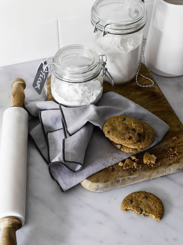 A styled baking scene on a pale marble countertop, featuring glass jars of flour, a white rolling pin, a folded grey kitchen towel, a wooden cutting board, and chocolate chip cookies. The image has a bright, clean, neutral farmhouse-inspired feel.