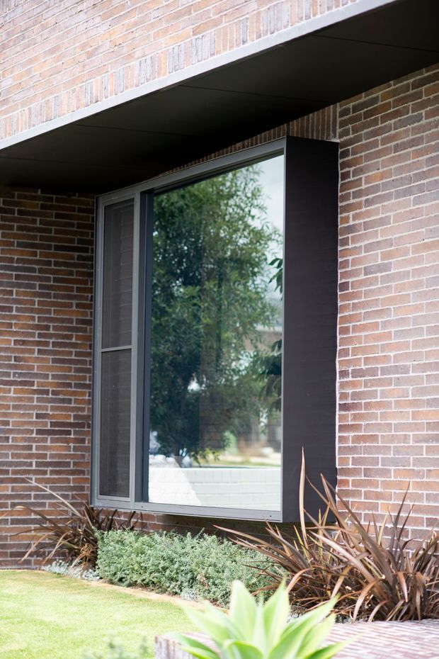 Exterior close-up of a contemporary brick facade with a large dark-framed window and adjacent screened opening beneath a deep projecting brick overhang. Low landscaped planting borders the base of the wall, and trees are reflected in the glazing.