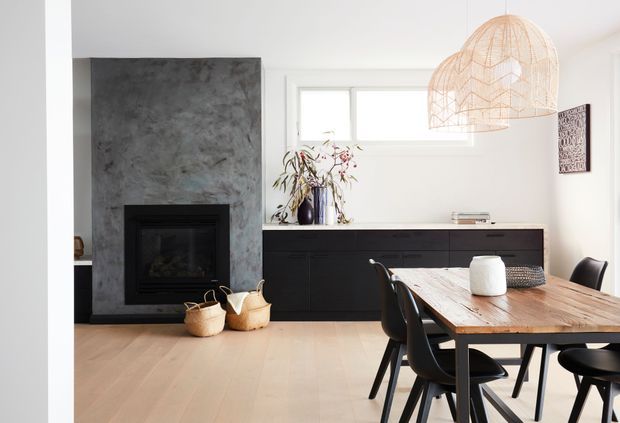 Bright contemporary dining area with a substantial timber dining table and black dining chairs in the foreground. A dark built-in sideboard runs along the rear wall beneath a high horizontal window, while a textured charcoal fireplace surround and black firebox form a strong feature at left. Two large woven pendant lights hang above the table, with pale timber flooring and white walls creating a light, warm interior.