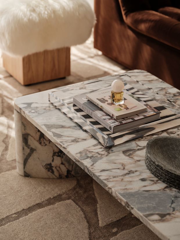Close-up interior vignette of a refined living room centered on a substantial rectangular marble coffee table with a rounded edge and matching marble-clad side support. A clear acrylic tray holds stacked design books and a small decorative object. A sculptural white boucle stool, brown leather seating, and a soft neutral patterned rug complete the warm, luxurious setting.