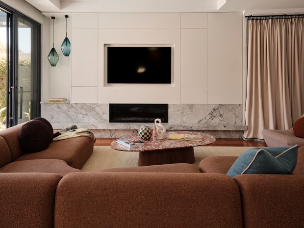 A refined living room viewed from behind a large brown sectional sofa. The room features a pale built-in media wall with a wall-mounted television, linear fireplace, and dramatic marble surround, along with floor-to-ceiling curtains, dark-framed glazing, sculptural pendant lights, and a low organic-shaped coffee table. The palette is warm and earthy with contemporary detailing.
