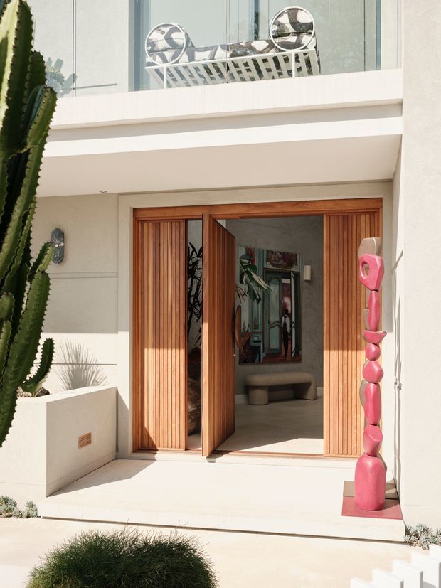Bright exterior view of a refined residential entryway with a pale rendered facade, broad balcony, and large vertical timber pivot doors. The entrance opens into a stylish interior with artwork and seating visible beyond. Sculptural pink decor and tall sculptural cacti flank the doorway, while a glass balcony balustrade and outdoor seating are visible above.