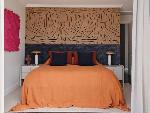 A styled bedroom viewed head-on, featuring a large bed with a vivid orange bedspread, layered navy and rust cushions, and a dark upholstered headboard. A bold abstract timber-toned graphic wall panel sits above the bed, flanked by bedside tables with sculptural lamps. The room has pale walls, white cabinetry, carpeted flooring, decorative wall art, and traditional ceiling cornices.