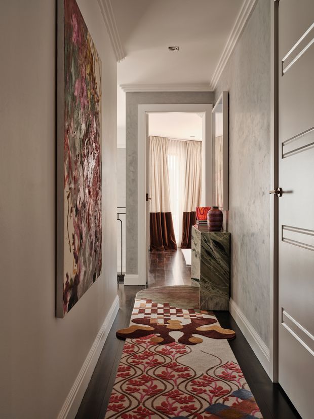 A refined residential hallway viewed toward a curtained room, with dark timber flooring, pale textured walls, ornate white crown moulding and skirting, paneled doors, and a narrow console featuring a dramatic green stone finish. Layered patterned runners, a large abstract floral artwork, sculptural ceramics, and warm drapery add a richly decorated, eclectic character.
