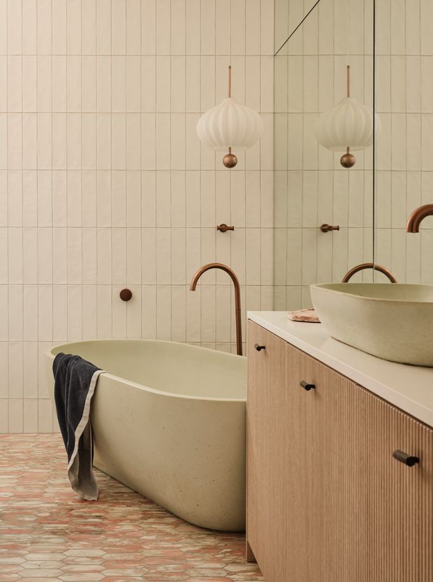 A warm, refined bathroom featuring a freestanding oval bathtub, wall-mounted copper-toned faucet, vessel basin, and fluted timber vanity cabinetry. The walls are clad in narrow cream vertical tiles, while the floor has a decorative pink and terracotta patterned tile finish. A large mirror reflects the pendant light and fittings, and sculptural cream pendant lighting adds a soft focal point.