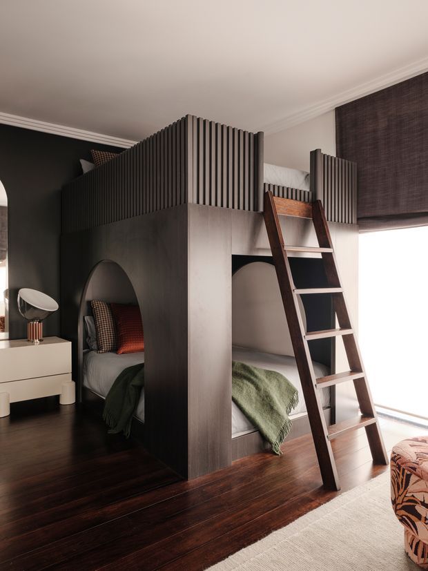 A stylish compact bedroom featuring a custom dark timber bunk-bed structure with arched openings, integrated upper bunks enclosed by vertical slatted screens, and a wooden ladder. The lower bunks have muted grey bedding with green throws and patterned cushions. The room includes dark polished timber flooring, a white bedside cabinet with a sculptural lamp, a large dark roller blind, and a patterned upholstered chair or ottoman in the foreground. The overall design is refined, contemporary, and space-efficient.