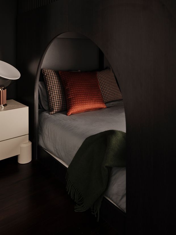 A moody, intimate bedroom viewed through a tall arched opening in dark timber-toned joinery or wall panelling. The bed is dressed in grey bedding with patterned cushions, a rust-red textured accent cushion, and a dark green throw. A minimalist white bedside cabinet, sculptural lamp, and small cylindrical waste bin sit to the left. Dark flooring and walls create a dramatic, cocooning atmosphere.