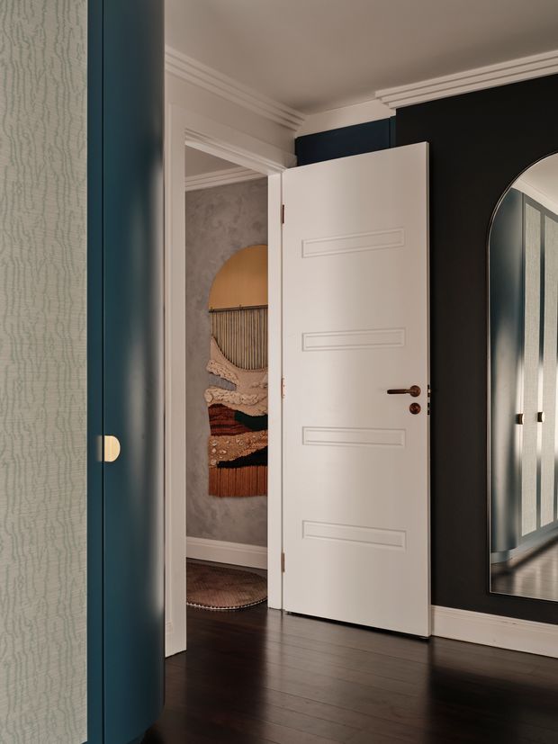 A refined residential hallway or entry area with a partially open white paneled door, dark timber flooring, deep navy-black walls, ornate white crown moulding, and a large arched mirror. The mirror reflects additional blue cabinetry or wall panelling and a bright adjoining space. A textured decorative wall hanging is visible beyond the doorway, and the space has a moody, elegant, layered interior design.