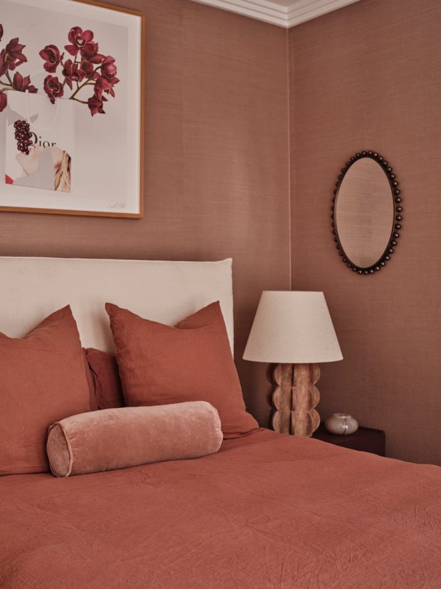 A warmly styled bedroom corner featuring a terracotta bedspread and matching pillows against textured terracotta walls. A white upholstered headboard, sculptural bedside lamp, oval beaded mirror, framed floral artwork, and dark bedside table create a refined, richly coloured interior.