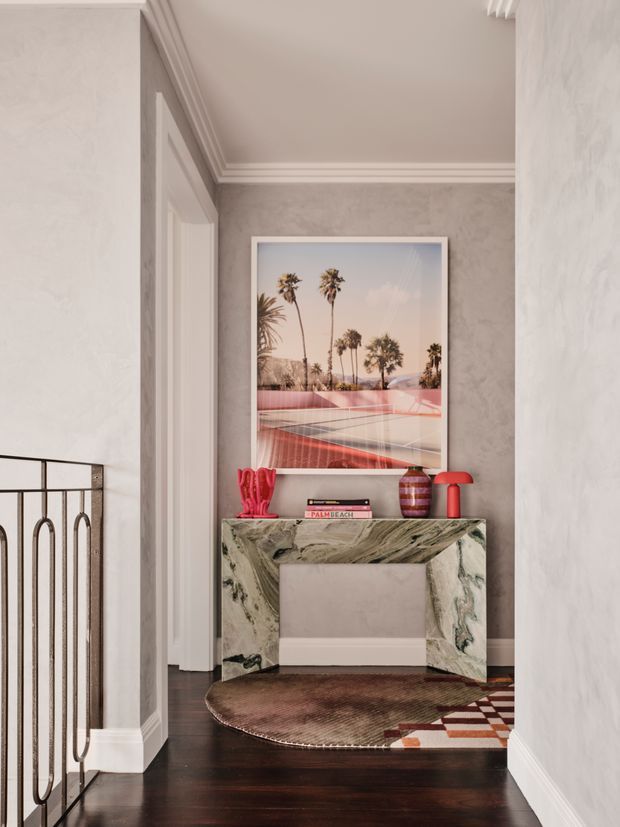 A refined residential hallway or landing with dark timber flooring, pale textured walls, ornate white crown moulding and door trim, and a sculptural veined-stone console table. A framed palm-tree artwork, colourful decorative objects, and layered patterned rugs create a playful yet sophisticated look. A stair balustrade is visible at left, confirming the circulation space.