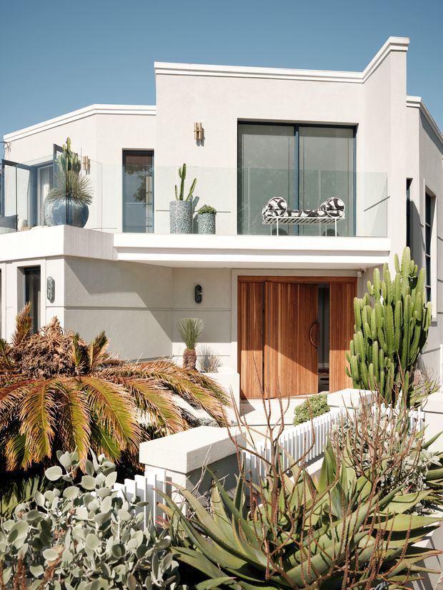 A bright exterior view of a two-storey contemporary residence with crisp white rendered walls, clean geometric rooflines, extensive dark-framed glazing, a glass-balustraded upper terrace, and a large warm timber pivot entry door. The property is surrounded by drought-tolerant succulent and cactus landscaping, with a sculptural bench on the balcony and strong midday sunlight.
