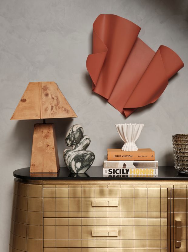 A styled interior vignette featuring a curved, grid-panelled brass-gold sideboard with dark stone-like top, set against a textured grey wall. The arrangement includes a warm timber table lamp, sculptural stone ornament, white pleated vase, stacked books, and a terracotta-red abstract wall sculpture. The overall look is refined, warm, and contemporary with luxe accents.