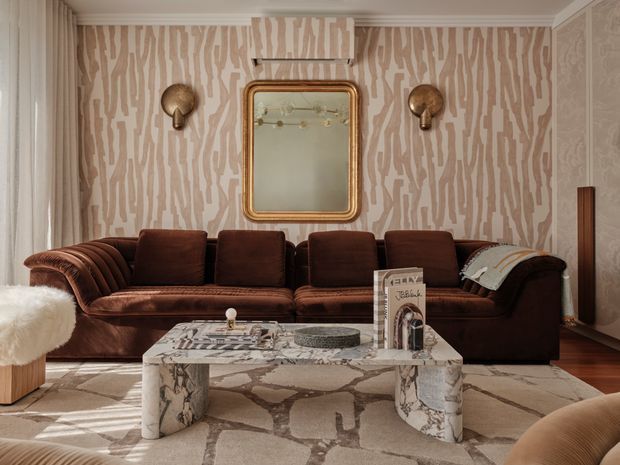 A richly styled living room with a deep brown velvet sectional sofa, patterned beige-and-taupe wallpaper, a large framed mirror, sculptural wall sconces, and a veined stone coffee table. A neutral patterned rug, warm timber flooring, and layered furnishings create a sophisticated, moody interior with strong Art Deco-inspired detailing.