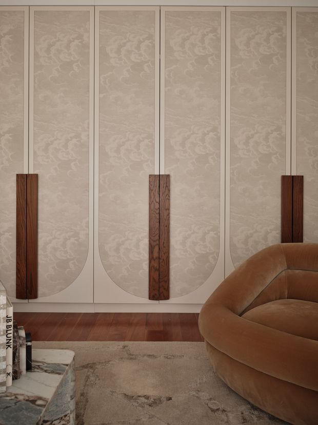 A refined, moody interior detail featuring a wall of tall decorative panels with pale textured patterned inserts and curved lower edges, accented by narrow dark timber handles or appliqué panels. A warm brown upholstered curved lounge chair occupies the foreground, with dark timber flooring, a neutral textured rug, and a stone coffee table partially visible. The composition emphasizes bespoke wall panelling, sculptural furniture, and a warm neutral palette.