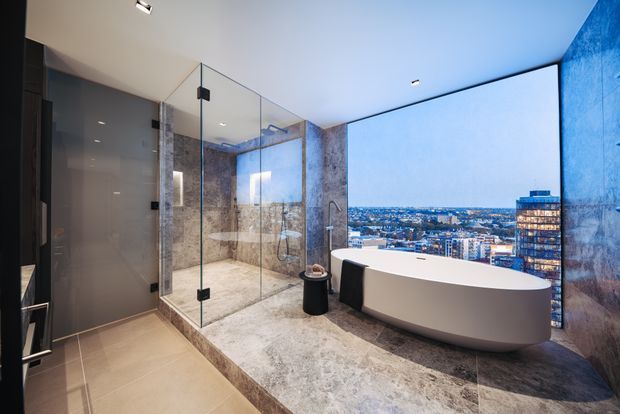 Luxurious contemporary bathroom with a freestanding white bathtub positioned beside floor-to-ceiling glazing overlooking a dense urban cityscape. The room includes a large frameless glass shower enclosure, stone-look wall and floor finishes, dark cabinetry or wall panels, recessed ceiling lighting, and a striking panoramic view.