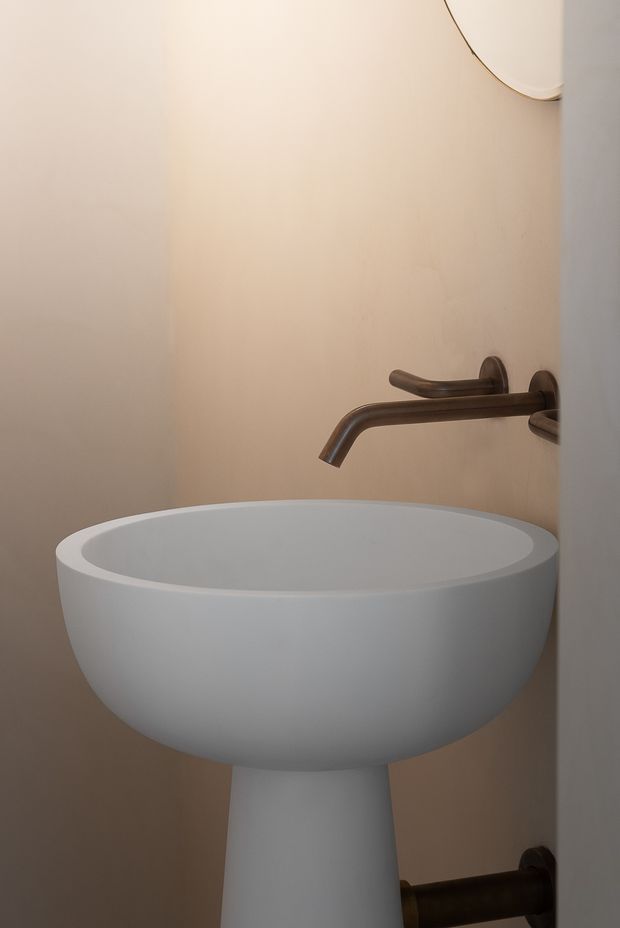 Close-up of a sculptural white pedestal basin in a compact bathroom, set against warm beige walls. A wall-mounted bronze-toned faucet and plumbing are visible, with part of a rounded mirror at the upper right. The space has a calm, minimalist, refined appearance.