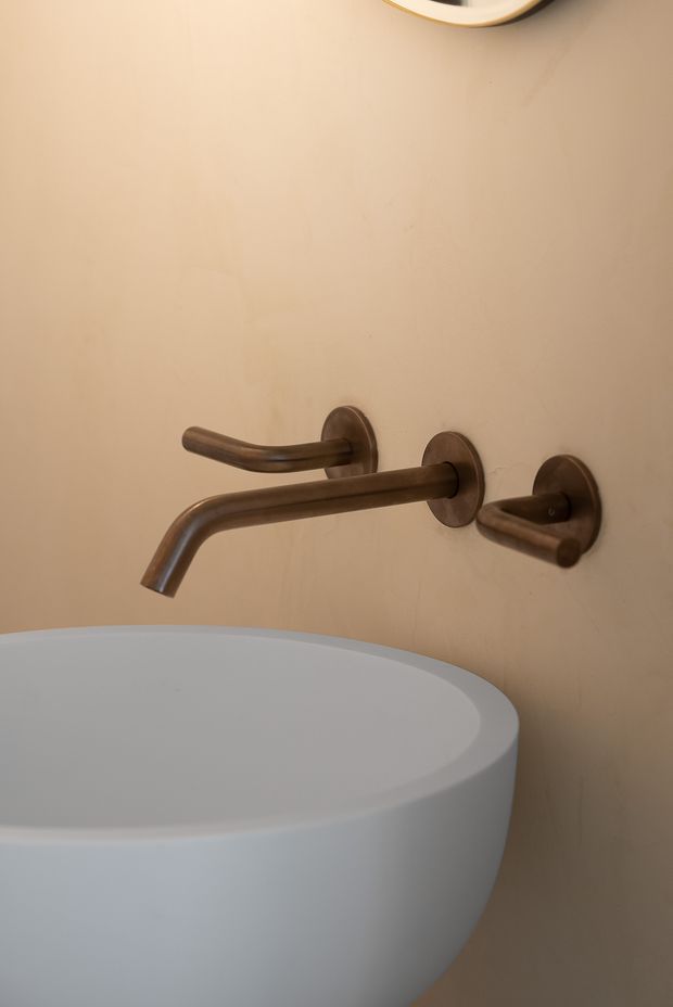 Close-up of a contemporary bathroom basin with a curved white vessel sink and wall-mounted brushed bronze/copper tapware against a warm beige plastered wall. A partial round mirror is visible at the top edge.