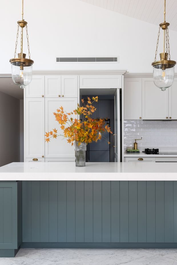 Bright, refined kitchen with a large island featuring blue-grey vertical-panel cabinetry and a white stone benchtop. The room has white shaker-style cabinetry, a glossy white subway-tile splashback, brass hardware and matching hanging lantern pendants, with pale marble-look flooring. A vase of orange foliage provides a warm accent at the centre of the island.
