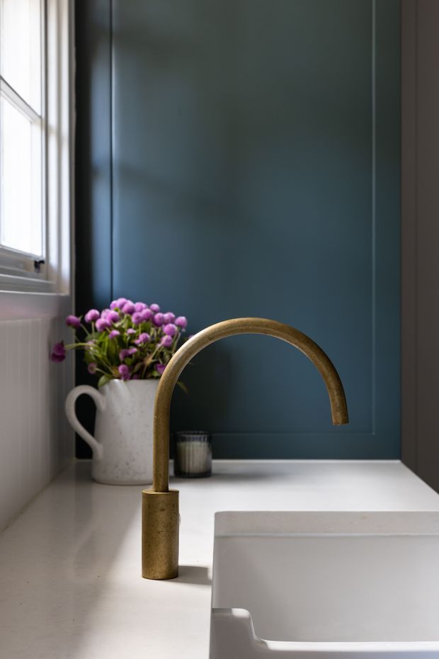 Close-up interior view of a refined bathroom vanity, featuring a sculptural brushed brass gooseneck tap, white integrated basin, pale stone or solid-surface countertop, dark blue-green wall panelling, and a white-framed window. A ceramic jug with purple flowers adds a decorative accent.
