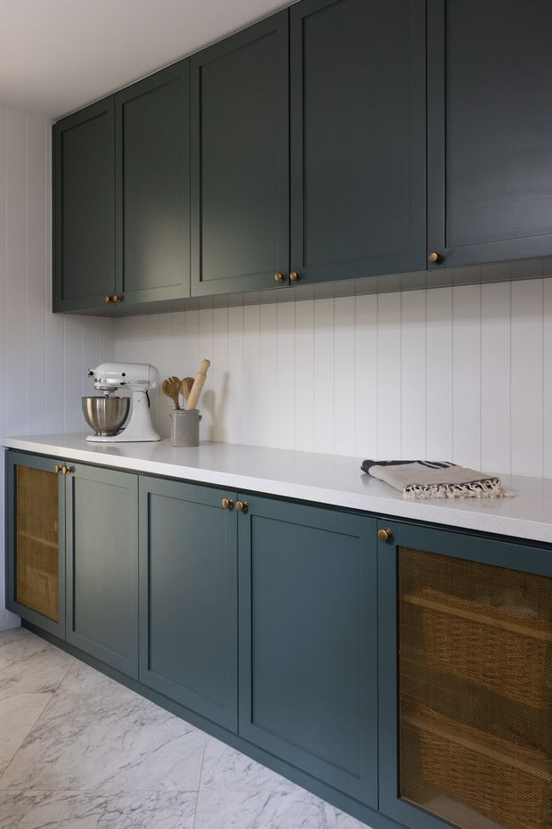 A refined kitchen or butler's pantry wall with deep blue-green framed cabinetry, brass knobs, a white countertop, and white vertically grooved wall panelling. A stand mixer and utensil crock sit on the counter, while the lower cabinetry includes a dark woven-mesh-front section. The floor is light marble-look tile, creating a classic yet contemporary appearance.