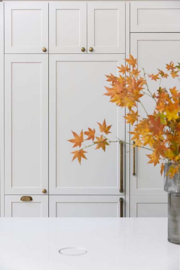 Close-up view of a light neutral built-in kitchen with classic recessed-panel cabinetry, slim dark reveals, and mixed brass and dark metal hardware. A white island or benchtop occupies the foreground, while a ribbed glass vase with autumn branches adds a warm decorative accent.
