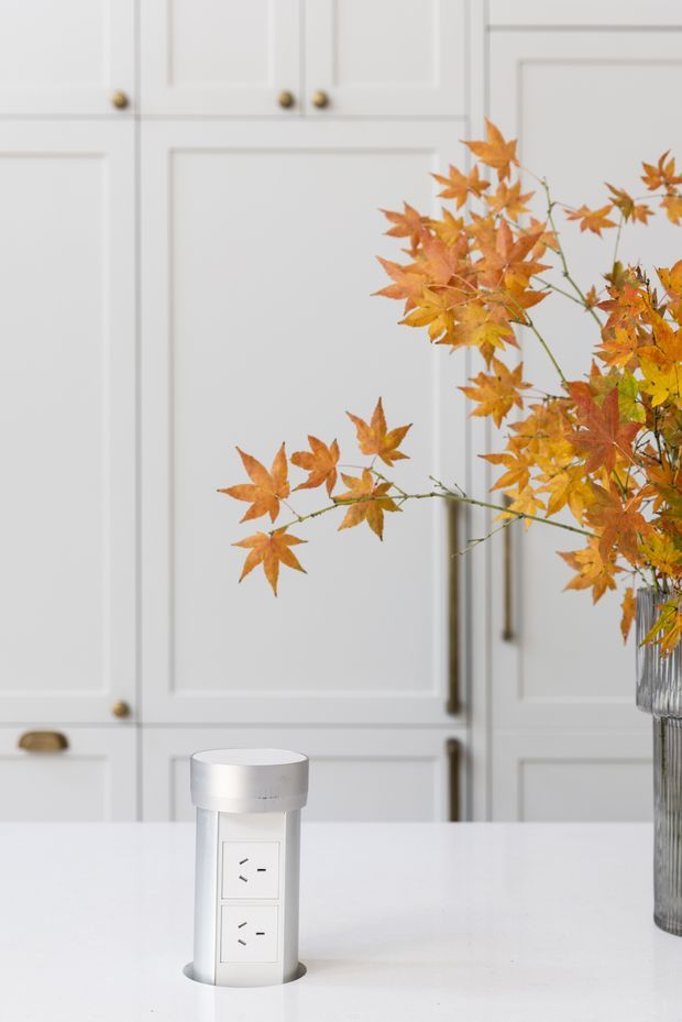 Bright, refined kitchen interior featuring white Shaker-style cabinetry with brass knobs and handles, a white stone island benchtop, and a cylindrical pop-up power outlet integrated into the island. A glass vase of orange autumn foliage provides a warm accent.