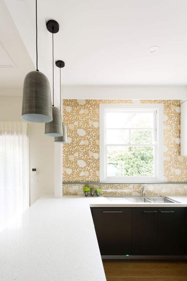 Bright contemporary kitchen with a large white peninsula benchtop, dark flat-panel cabinetry, stainless-steel sink, patterned botanical wallpaper, and three oversized dark pendant lights. A white-trimmed window brings in abundant natural light, with sheer curtains and timber flooring visible.