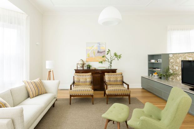 Bright, inviting living room with a pale neutral palette, timber flooring, a large area rug, cream sofa, patterned mid-century-style armchairs, green lounge chairs, and a compact media cabinet. Decorative wall panelling, framed artwork, indoor plants, sheer curtains, and a sculptural pendant add warmth and character.