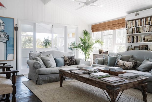 Bright coastal-style living room with white vertical wall panelling and a white-painted timber ceiling, two slipcovered sofas, a large distressed wood coffee table, woven rug, indoor palms, woven window blinds, and built-in open shelving filled with books. Large windows provide abundant natural light and views of palm trees and nearby buildings.