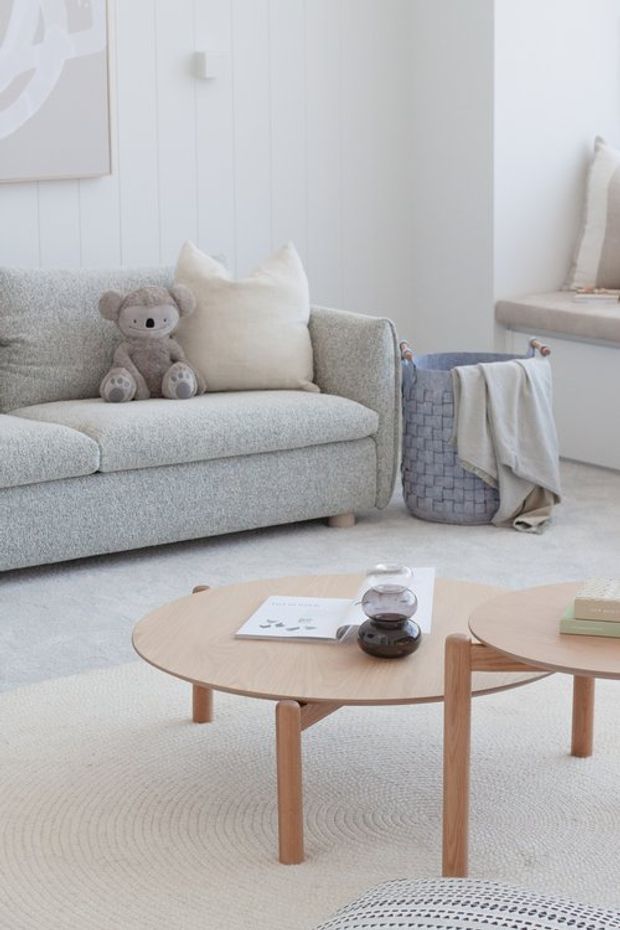 A bright, softly styled living room with a pale grey upholstered sofa, neutral cushions, a plush light rug, and two light timber round coffee tables. A woven basket with folded throws sits beside the sofa, while a window seat and white vertical wall panelling reinforce the airy, minimalist Scandinavian feel. A plush koala toy adds a subtle child-friendly touch.
