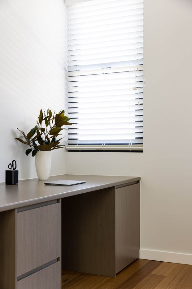 A bright, compact home office featuring a built-in L-shaped timber-look desk with integrated drawers and cabinetry, positioned beneath a large window with white horizontal blinds. The room has white walls, warm timber flooring, a simple potted plant, and minimal desk accessories, creating a clean contemporary workspace.