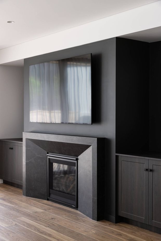 Contemporary living room fireplace feature wall with a dark grey stone surround, black built-in cabinetry, a wall-mounted television, and light timber flooring. The room has a restrained monochromatic palette with clean, streamlined detailing.